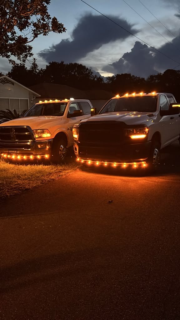 Dos camionetas RAM blancas de trabajo pesado estacionadas al atardecer con luces ámbar encendidas.