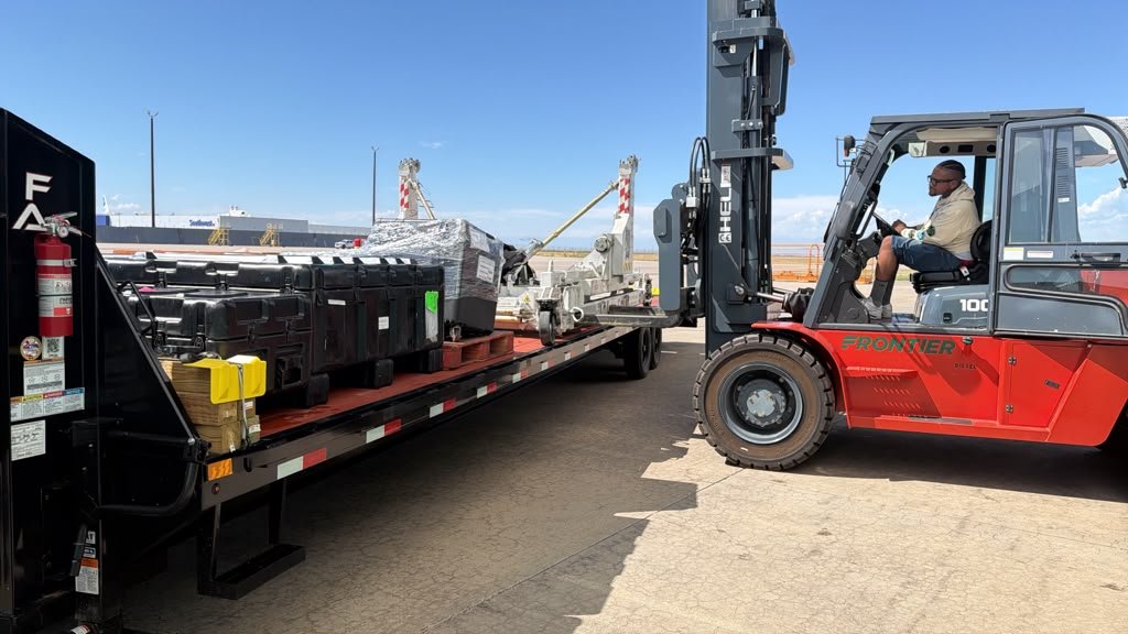 Operador de montacargas junto a un tráiler de plataforma con equipos industriales durante una operación de carga.