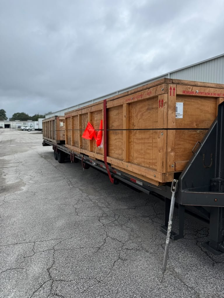 Tráiler gooseneck de plataforma transportando grandes cajas industriales de madera con correas de sujeción y banderas rojas.