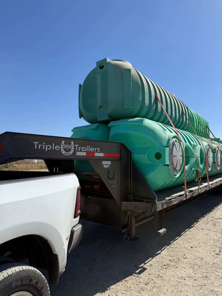Pickup blanca con tráiler gooseneck transportando grandes tanques plásticos verdes asegurados con correas.Pickup blanca con tráiler gooseneck transportando grandes tanques plásticos verdes asegurados con correas.